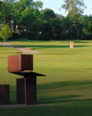 Escultura moderna de metal en un parque verde con árboles al fondo y césped amplio