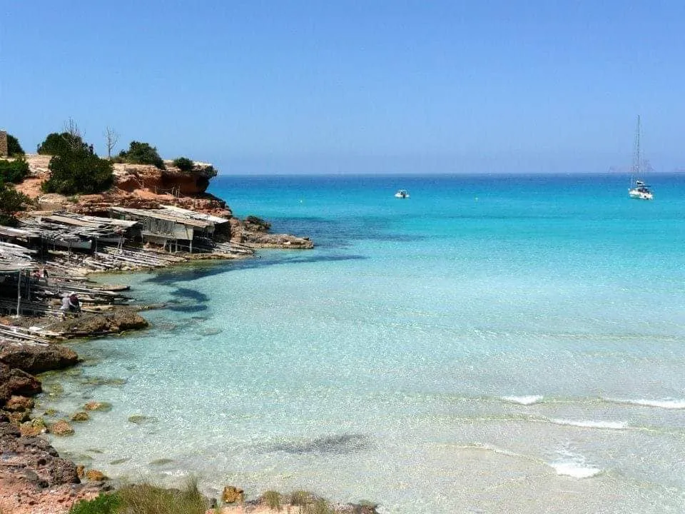 formentera-playa-cristalina Playa de una cala paradisiaca de con agua cristalina de Formentera con veleros en el fondo y un día soleado sin nubes.