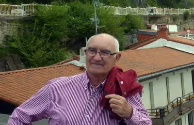 jesus-mari-alquezar Hombre mayor con camisa a rayas y chaqueta en el hombro frente a edificios y vegetación